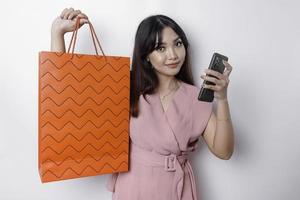 Portrait Asian happy beautiful young woman standing excited holding an online shopping bag and her smartphone, studio shot isolated on white background photo