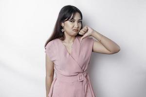 Disappointed Asian woman wearing pink blouse gives thumbs down hand gesture of disapproval, isolated by white background photo