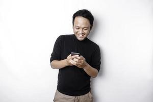 emocionado asiático hombre vistiendo negro camisa sonriente mientras participación su teléfono, aislado por blanco antecedentes foto