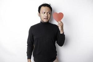 A portrait of an Asian man wearing a black shirt looks so confused while holding a red heart-shaped paper, isolated by a white background photo