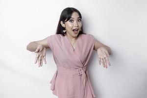 Shocked Asian woman dressed in pink, pointing at the copy space below her, isolated by white background photo