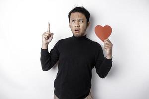 Confused Asian man holding a big red heart symbol pointing up at copy space isolated over white background photo