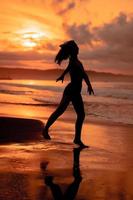 silueta de un asiático mujer practicando su ballet se mueve en el playa con el olas estrellarse antes de el festival empieza foto