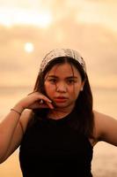 an Asian teenager wearing a white bandana and black shirt with a flat expression while posing with her hair on the beach photo