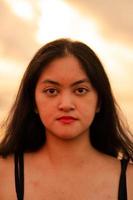 An Asian woman in a black shirt with an expression that looks annoyed is standing near the waves on the beach in a holiday photo