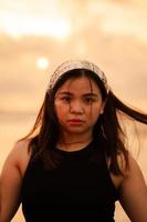 an Asian teenager wearing a white bandana and black shirt with a flat expression while posing with her hair on the beach photo