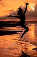 un balinés mujer en el formar de un silueta realiza ballet movimientos muy hábilmente y flexiblemente en el playa con el olas estrellarse foto