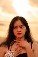 An Asian woman with black hair and a beautiful face posing in front of the beach with warm photo