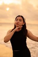 a glamorous Asian woman in black clothes and a white bandana standing in front of the waves on the beach photo