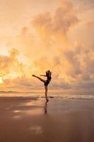 A ballerina in a black dress is practicing ballet moves on the beach with very flexible movements with a view of the clouds behind photo