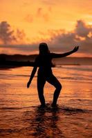 un balinés mujer en el formar de un silueta realiza ballet movimientos muy hábilmente y flexiblemente en el playa con el olas estrellarse foto