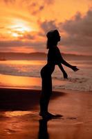 silueta de un asiático mujer practicando su ballet se mueve en el playa con el olas estrellarse antes de el festival empieza foto