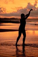 un balinés mujer en el formar de un silueta realiza ballet movimientos muy hábilmente y flexiblemente en el playa con el olas estrellarse foto