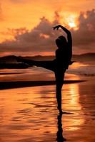 un balinés mujer en el formar de un silueta realiza ballet movimientos muy hábilmente y flexiblemente en el playa con el olas estrellarse foto