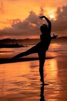 un balinés mujer en el formar de un silueta realiza ballet movimientos muy hábilmente y flexiblemente en el playa con el olas estrellarse foto