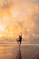 An Asian woman in a black dress performs ballet movements beautifully on the beach with waves and clouds behind her photo