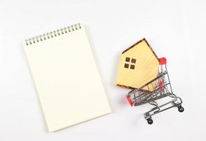 flat layout of wooden house model in shopping trolley or shopping cart with blank page opened notebook on white background, home purchase concept. photo