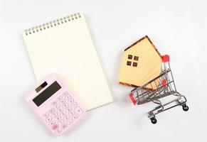 flat layout of wooden house model in shopping trolley, pink calculator, blank page opened  notebook  on white background with copy space, home purchase concept. photo