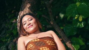 An Asian woman is sleeping on a tree trunk full of green leaves while enjoying the calming island's atmosphere video