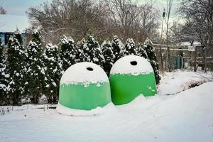 Green round plastic dumpster waste and garbage containers on snow in winter season near fir trees fence photo