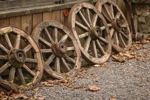Old wooden wheels arranged in a row near wooden plank elevation on pebble road with autumn leaves photo