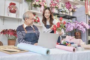 Asian senior male florist owner discusses with young beautiful female employee about bunch of fresh blossom arrangements for online business, happy work in colorful flower shop store, e-commerce SME. photo