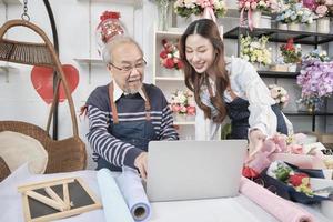 Asian elder male florist owner discussing with young beautiful female employee who shopkeeper about website arrangement for business service, happy work in colorful flower shop store, e-commerce SME. photo