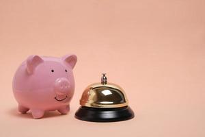 Piggy bank and gilded bell hotel service on pastel beige background.Conceptual, expensive hotel, travel and recreation.