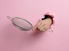 metal sieve for sifting flour in a female hand on a pink background. Part of the body sticking out of a torn hole in a paper background photo