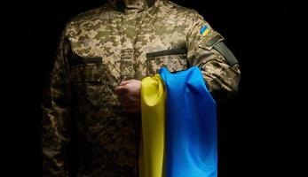un soldado de el ucranio armado efectivo soportes con un azul amarillo bandera de Ucrania en un negro antecedentes. honrando veteranos y conmemorando esos delicado en el guerra foto