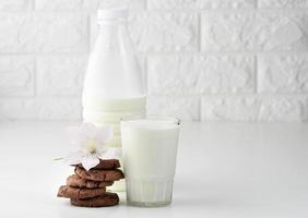 a full glass cup of milk and a clear plastic bottle with milk, next to a stack of round chocolate chip cookies on a white table photo