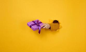 female hand holds a box with a gift on a yellow background, part of the body sticks out of a torn hole in a paper background. Congratulation, holiday surprise photo