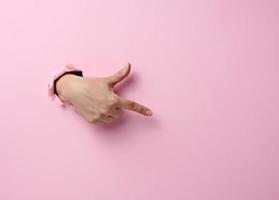 female hand sticks out of a torn hole in a pink paper background, part of the body points with the index finger to the side photo