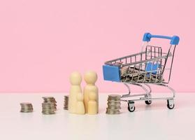 wooden figurines of a family and stacks of metallic money, a miniature shopping cart on a white table. Family budget control concept, savings photo