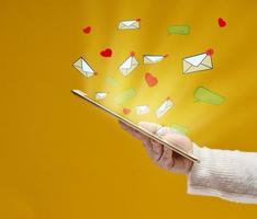 female hand holds a tablet with flying out hearts, dialogues and letters on a yellow background. Social media addiction, receiving correspondence and appraisal. Multitasking photo