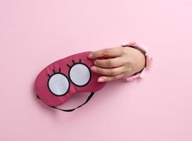 pink textile sleep mask in a female hand sticking out of a bull in a pink paper background, insomnia photo