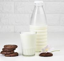 a full glass cup of milk and a clear plastic bottle with milk, next to a stack of round chocolate chip cookies on a white table photo