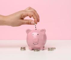 female hand throws a coin into a pink piggy bank on a white table. Concept of accumulating cash, saving, receiving subsidies photo