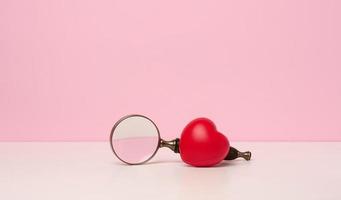 red heart and magnifier on white table, pink background photo