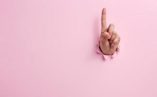 female hand sticking out of a torn hole in a pink paper background, attention gesture photo