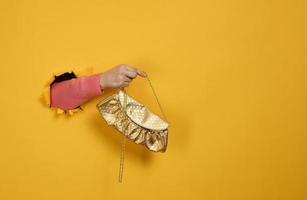 female hand is holding a small golden clutch bag with a metal chain on a yellow background. Part of the body sticking out of a torn hole in a paper background photo