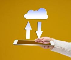 female hand holds an electronic gadget tablet on a yellow background, white cloud with arrows above it. Information exchange, data warehouse. Uploading a backup photo