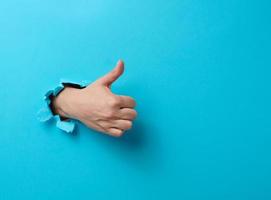 woman's hand sticks out of the torn hole and shows a gesture okay, thumb up. Blue background photo