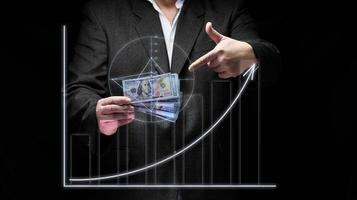 a businessman in a suit holds a pack of paper american dollars against the background of a holographic chart with growing indicators. The concept of high business profitability, income growth photo