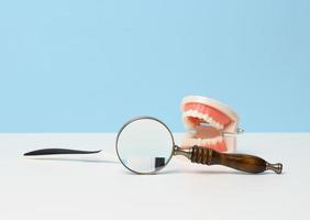 Magnifier, toothbrush and a plastic model of a human jaw with white even teeth and a medical examination mirror on a white table. Morning teeth cleaning, oral hygiene photo