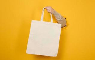 female hand holds an empty white textile bag on a yellow background, a part of the body sticks out of a torn hole in a paper background. Reusable and recyclable packaging, no plastic photo