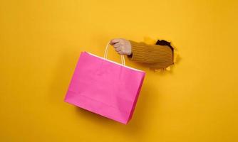 female hand with a pink paper shopping bag sticking out of a torn hole in a yellow paper background. Refusal from plastic, start of sales photo