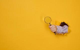 a female hand holds a magnifying glass on a yellow background, part of the body sticks out of a torn hole in a paper background. Business concept, finding information photo