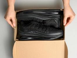 female hands are holding a brown cardboard box with black sports sneakers on a gray table, inspecting an online order, top view photo