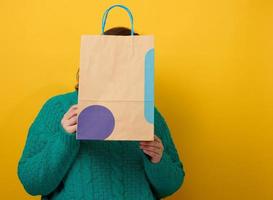 una mujer con suéter sostiene una bolsa de papel sobre un fondo amarillo y muestra un gesto similar con la mano. compras venta de temporada foto